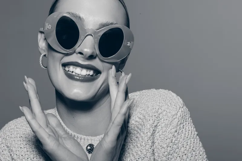 Happy smiling white woman with pale skin wearing round sunglasses and textured sweater poses with hands near face. Studio portrait with positive mood and fashion concept in monochrome style.