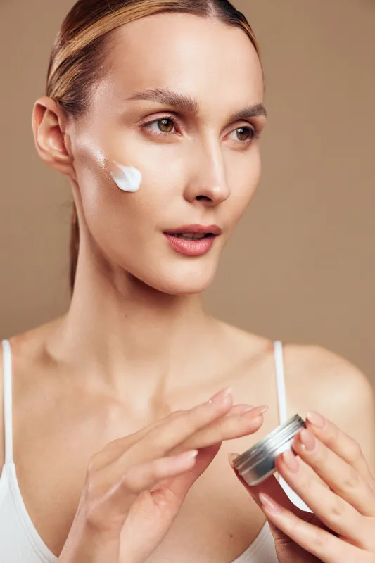 Young woman applies moisturizing cream on her cheek with fingers holds jar of skincare product looks focused isolated on beige background in beauty and wellness concept