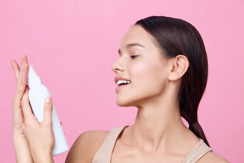 Young woman applying skincare product on her face, smiling and enjoying her routine against a pink background, showcasing beauty and self care