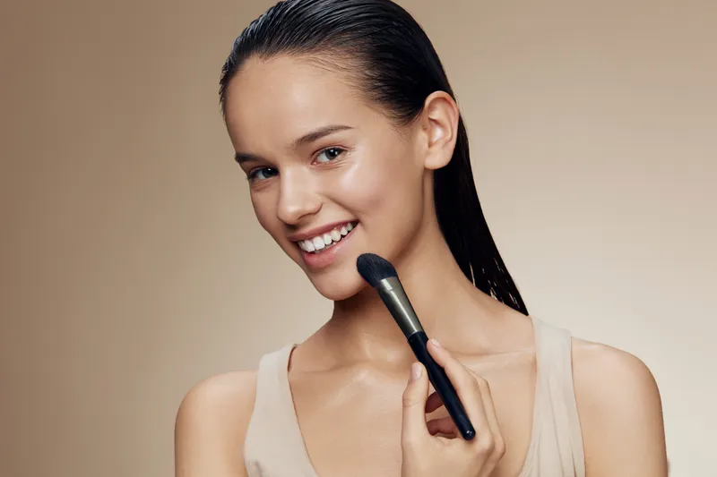 Young woman applying makeup with a brush, showcasing a natural beauty look, warm skin tones, and a simple background enhancing her radiant smile