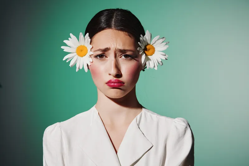Young woman with two daisies in her hair wears white clothing and pouty lips against a vibrant green background. Concept of fashion, beauty creativity, and tender emotions.