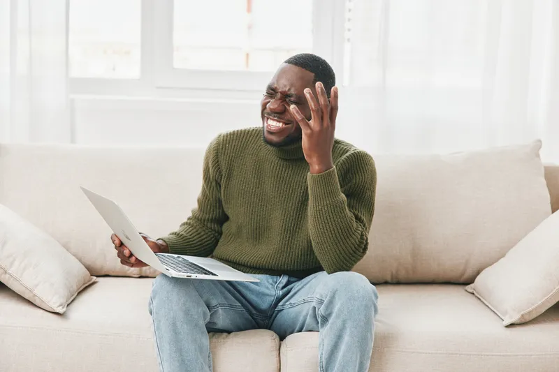 Young African man with laptop expresses frustration while sitting on a sofa, wearing a green sweater, highlighting a home office ambiance