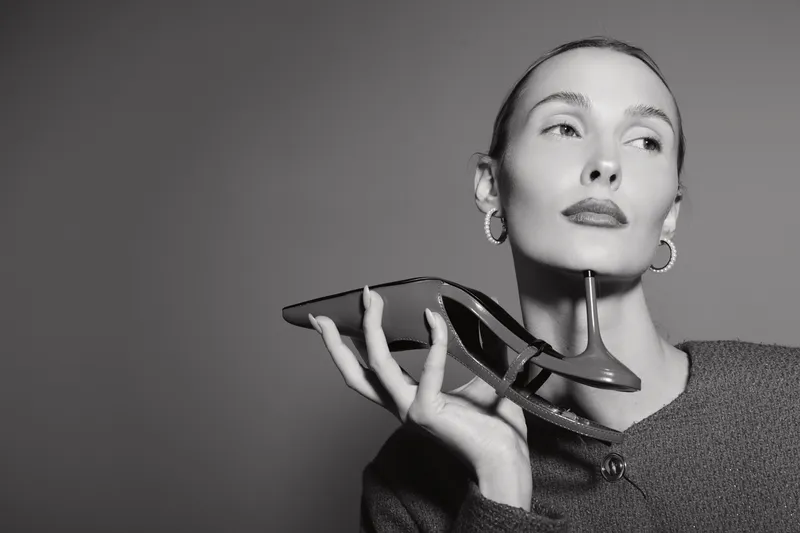 Young white woman holds high heel shoe near face looking away with confident expression. Concept for fashion, style, and elegance in monochrome photo.