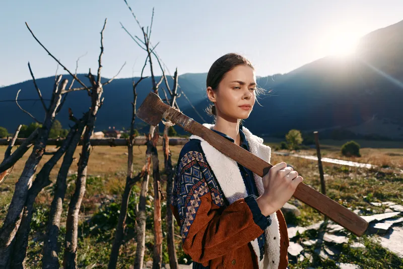 Woman with axe on her shoulder in a countryside farm setting by mountains at sunrise, portrait of a young rural worker in sweater carrying tool outdoors in golden sunlight.
