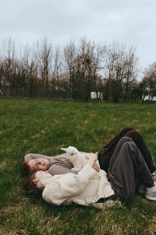 Relaxed person lying on grass with a friendly goat kid, enjoying a calm outdoor moment in a park, wearing a cozy sweater, surrounded by trees and a peaceful mood, daylight soft