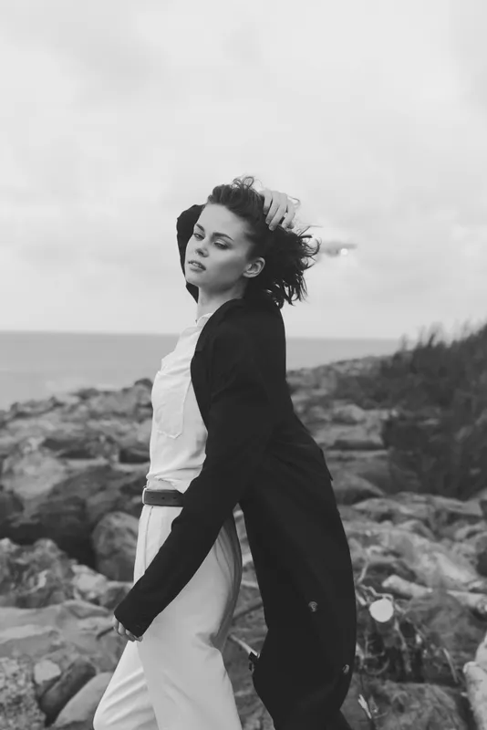 Monochrome fashion scene with a wind swept model by a rocky coast, showcasing an elegant coat and relaxed pose, outdoor seaside mood with dramatic texture and cinematic style