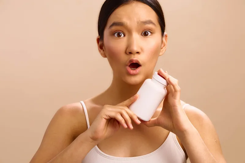 asian woman holds a white bottle with a surprised expression, skincare product in hand, casual top against a neutral background, creating a curious, approachable mood and highlighting everyday beauty
