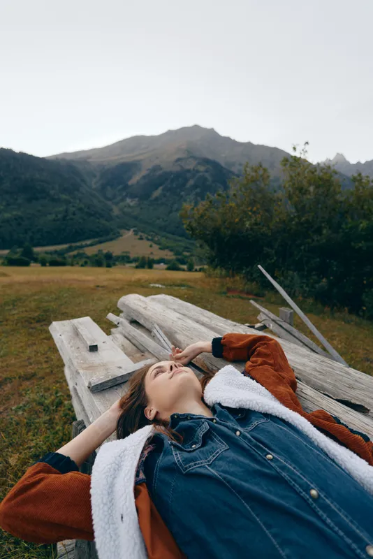 Woman portrait lying on a wooden bench in a meadow with mountains in the background, relaxation in nature and outdoors. Casual denim shirt and jacket, peaceful landscape escape.