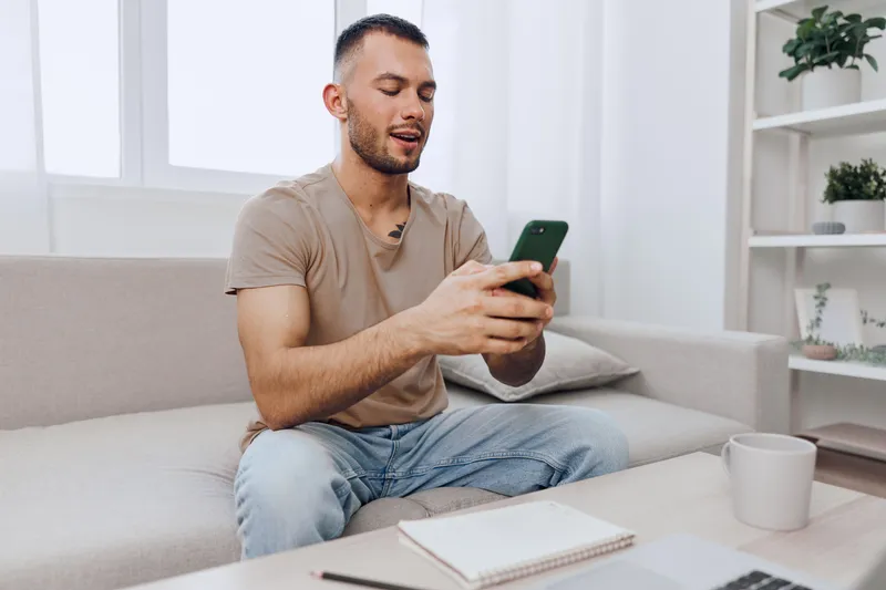 A focused man engaging with his smartphone, surrounded by a modern living space. Relaxed atmosphere, emphasizing technology and connectivity.