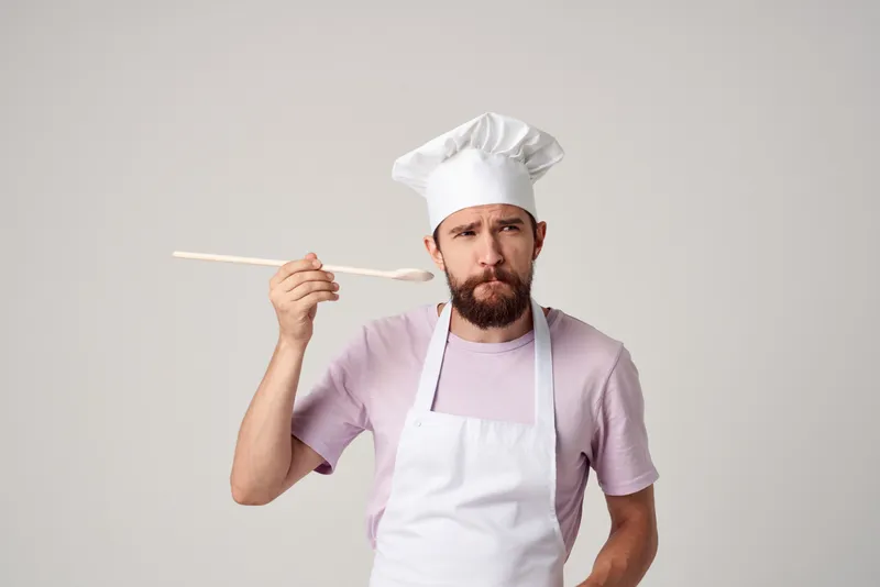 Повар-мужчина в колпаке пробует еду на вкус, занимаясь профессиональным приготовлением блюд