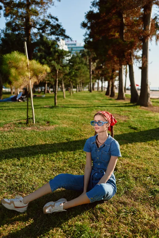 Lifestyle woman in modern boho-western denim street style sits relaxed on green grass, wearing bandana and sunglasses with film color tones and natural outdoor light.
