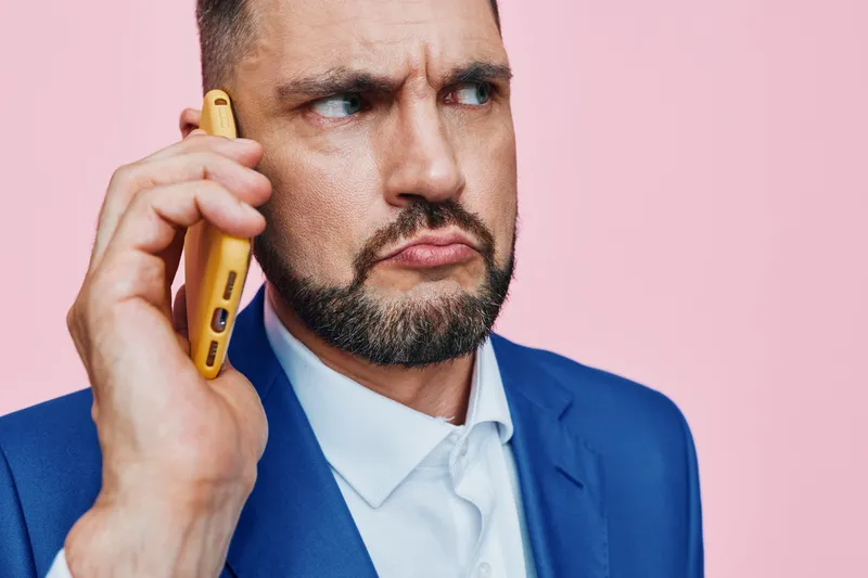 A focused business professional speaks on a mobile device against a solid pink backdrop, conveying determination, concentration, and seriousness in a high stakes moment.