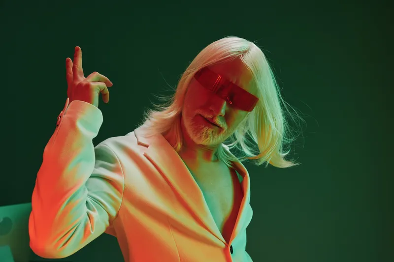 На этом выразительном изображении представлен мужчина с длинными седыми волосами и красными солнцезащитными очками в элегантном белом костюме