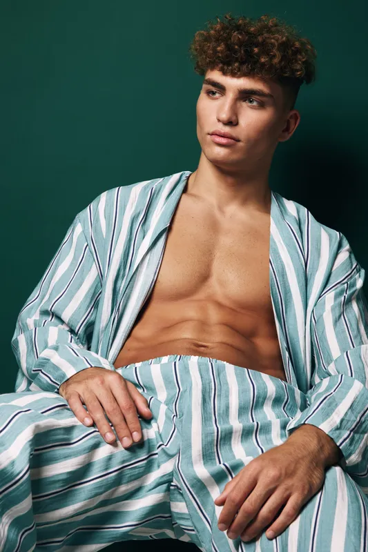 Young man with curly hair wearing striped open shirt and matching pants sitting relaxed against dark green background. Casual fashion, confident pose and toned body visible.