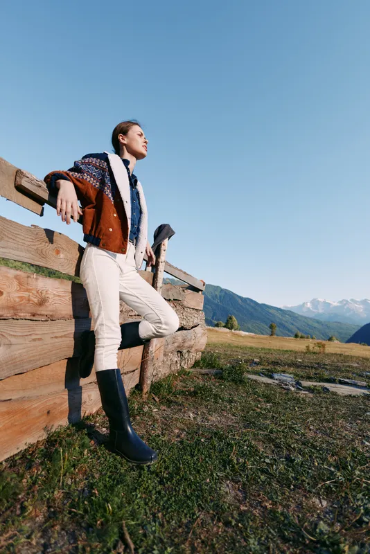 Woman leaning on wooden fence in countryside mountains, portrait outdoors wearing jacket and tall boots, looking into the distance during travel and nature lifestyle.