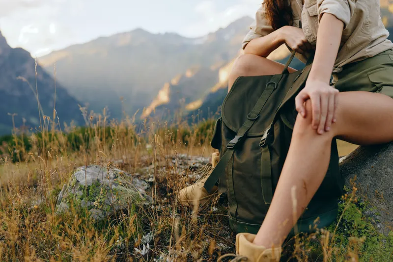 A woman sitting during a hiking moment in a sunlit mountain scenery, wearing casual clothing with a backpack nearby. Emphasizes outdoor lifestyle, nature, and calm.