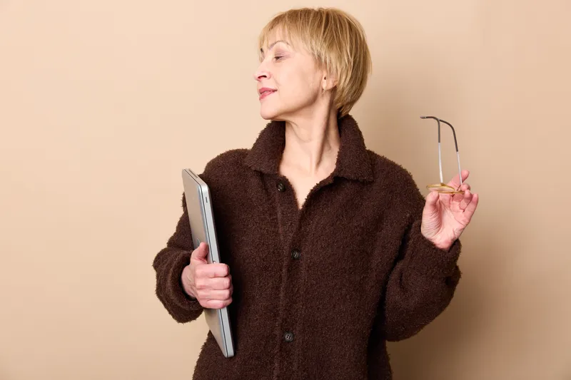 Woman with smartphone and headset stands in beige background, casual portrait, listening to audio, thoughtful expression, technology communication concept, modern life