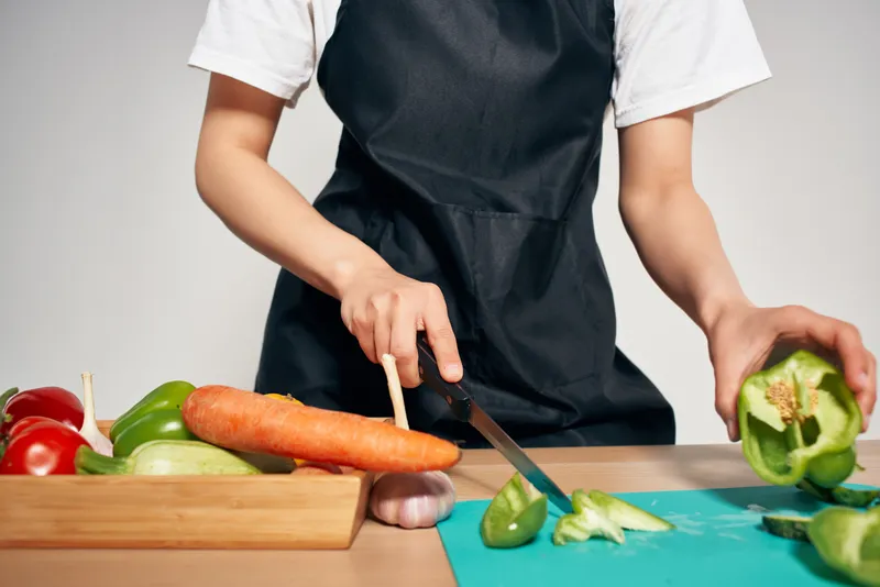 Женщина в черном фартуке нарезает овощи на кухне, готовит еду