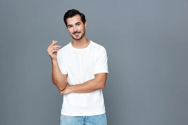 Relaxed young smiling man wearing casual white t-shirt stands confidently against a gray background, holding a hand up with a friendly expression. Modern lifestyle concept.