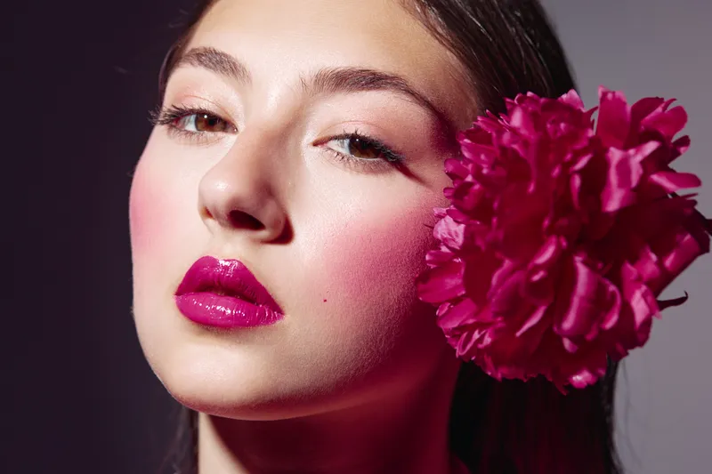 Portrait of a young woman with vibrant glossy fuchsia lips and pink cheeks wearing bold pink eyeshadow, paired with a large bright pink flower by her face for beauty and fashion concept.
