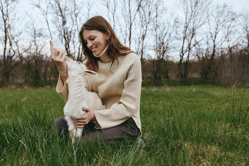 Smiling woman in a cozy sweater sits in tall grass with a playful goat kid, sharing a gentle outdoor moment. Natural light, serene countryside setting, bond between human and animal, warm mood,