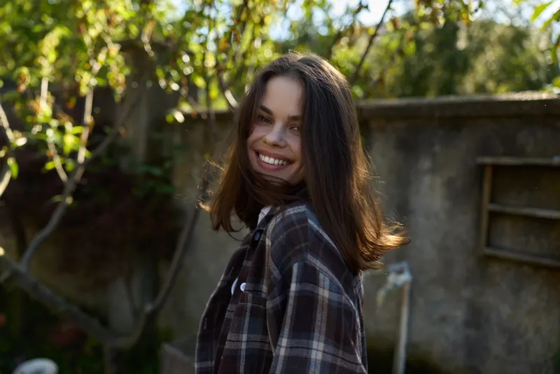 Young woman with shoulder length hair wearing a plaid shirt smiles softly in a sunlit garden, creating a natural lifestyle portrait with warm and welcoming vibes