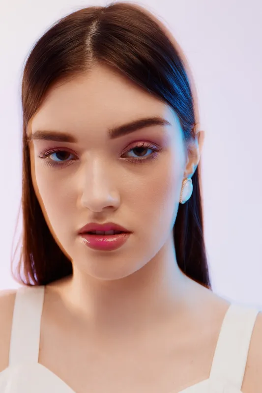 Close-up portrait of a young woman with smooth skin and glossy lips wearing minimal makeup and earrings. Studio shot with soft lighting. Concept of beauty, fashion, and elegance.
