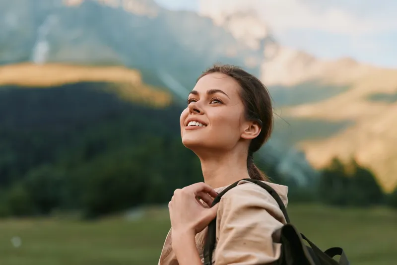 A woman outdoors with a backpack smiles in a sunny landscape, embodying travel and wellness. A wide mountain meadow and blue sky form a hopeful setting for lifestyle exploration.