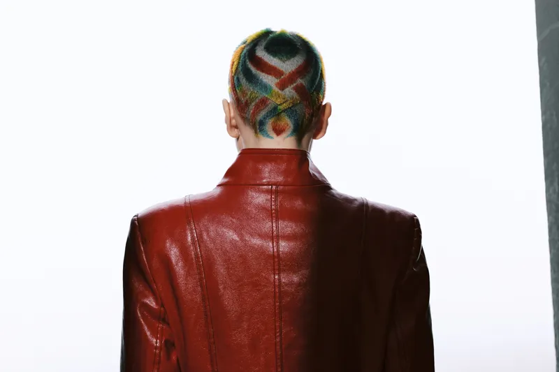 Woman with colorful shaved hairstyle wearing shiny red leather jacket, viewed from behind against white background, showcasing modern fashion and bold hair art.