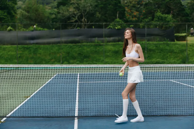 Young woman in sporty outfit on a tennis court, holding a tennis ball, showcasing athleticism and joy, with a vibrant green background enhancing the energetic atmosphere