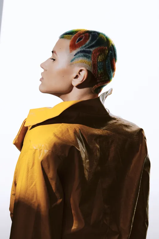Woman with colorful patterned shaved hair wearing a mustard yellow jacket, seen from side profile against a bright white background, showcasing artistic hairstyle and fashion.