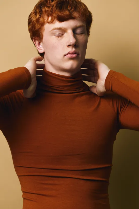 Young man with red hair wearing brown turtleneck sweater closes eyes and holds neck in contemplative pose against beige background for fashion or portrait concept.