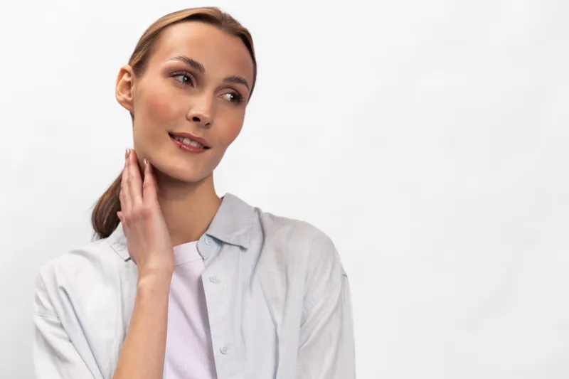 Young woman in casual white shirt touches her neck thoughtfully looking aside on white background. Concept of contemplation, beauty, natural expression, and casual lifestyle portrait.