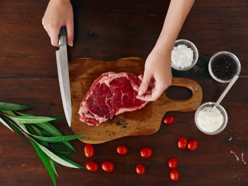 Женщина с ножом в руке нарезает свежее стейковое мясо для жарки на кухне