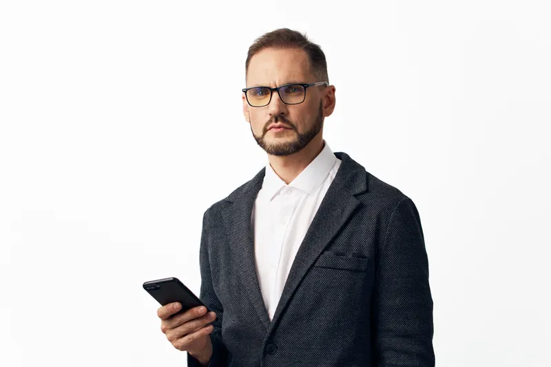 A confident business professional man stands against an isolated white background, holding a smartphone. He conveys focus, determination, and a poised executive demeanor.