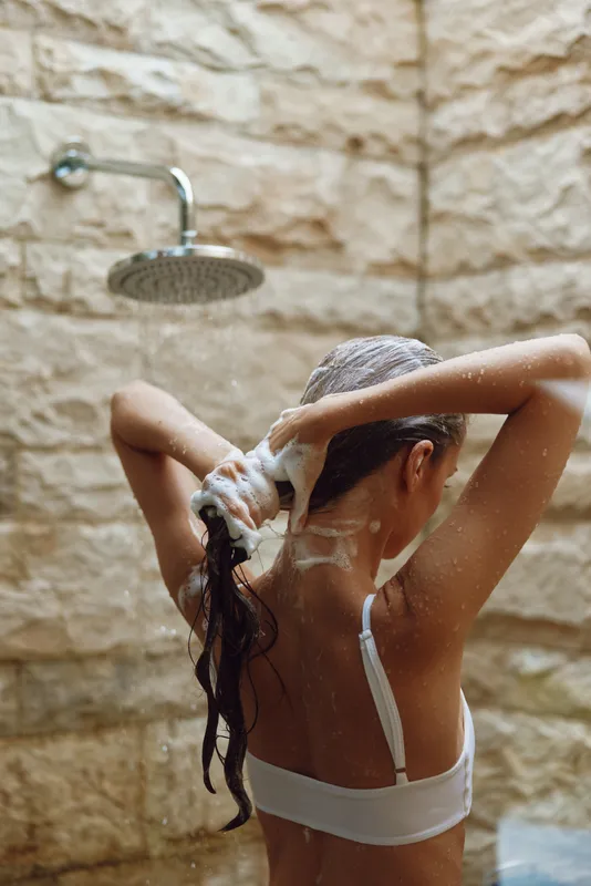 Woman washing hair with shampoo under outdoor rain shower, natural stone wall background, morning light, fresh lifestyle and self-care moment.