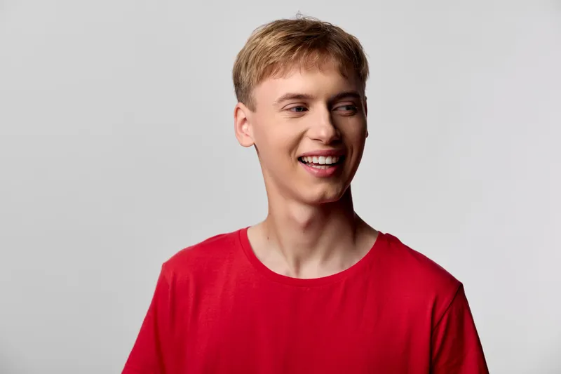 Smiling man in a red shirt stands against a light grey backdrop, casual style and friendly expression convey approachability and optimism, suitable for lifestyle, fashion or positive messaging