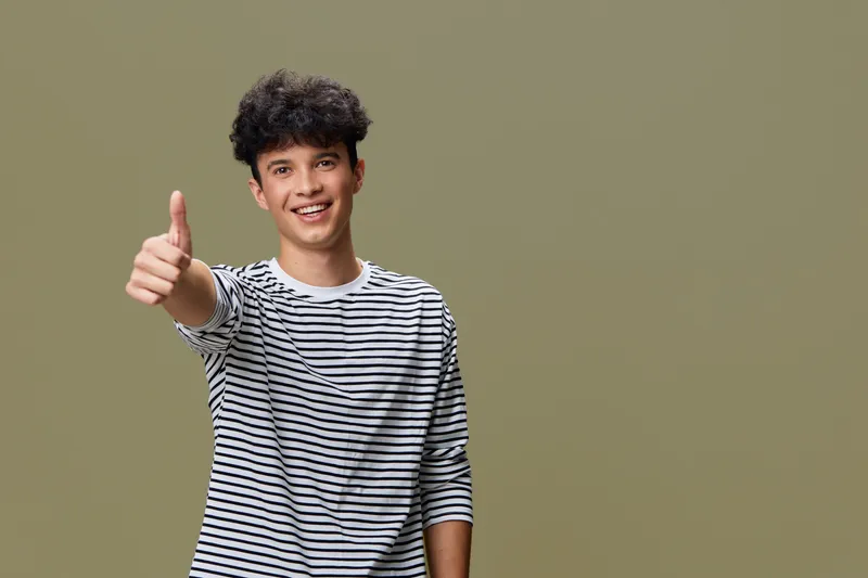 Young man with curly hair wearing striped shirt showing thumbs up gesture, smiling confidently against a neutral background in casual style.