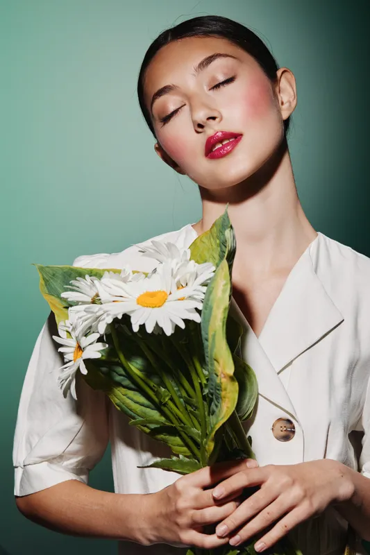 Young woman with radiant makeup and glossy red lipstick holds white daisies wrapped in green leaves. She wears a white blazer with closed eyes, posing against a soft green background.