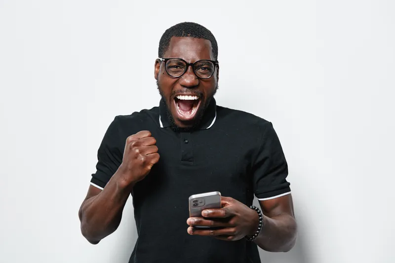 Excited African American man with dark skin wearing glasses and black polo shirt holding smartphone and celebrating success with fist pump on white background in studio.