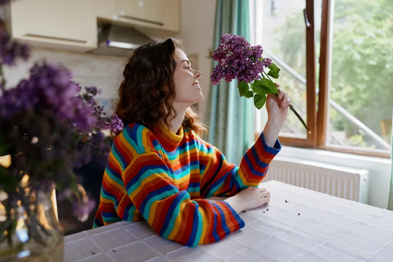Smiling woman with curly hair wearing a striped colorful sweater sits at a home table, admiring blooming flowers by a window, creating a cozy lifestyle scene with warm daylight.