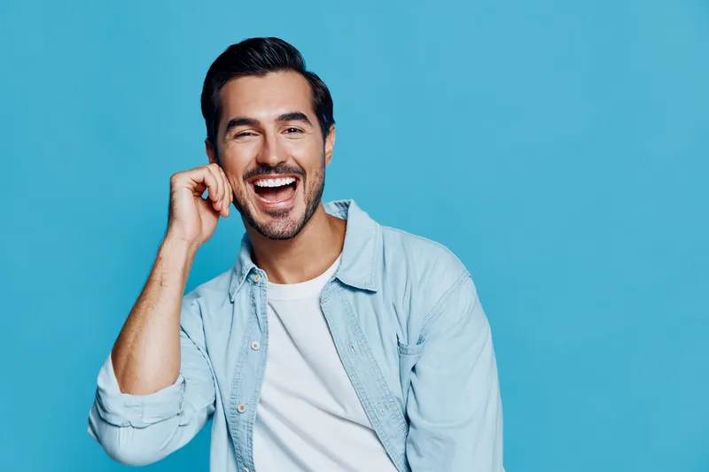 Smiling man with dark hair wearing a light blue shirt, joyfully posing with his hand on his chin against a vibrant blue background, conveying happiness and positivity.