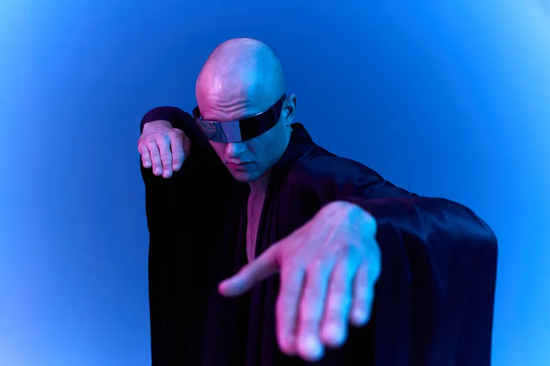 Futuristic man in shades striking a dynamic pose, dressed in a dark cloak against an ethereal blue background, creating a bold and captivating black and white image.