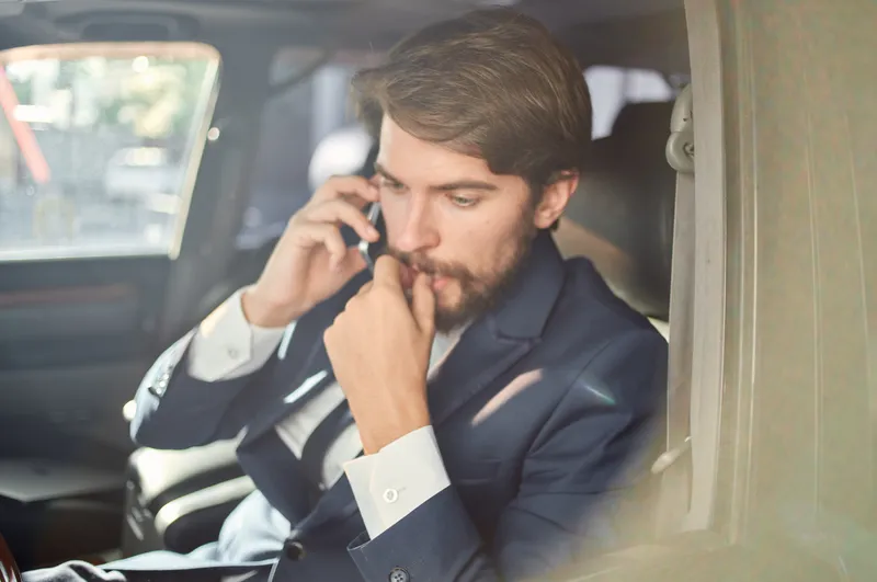Бородатый мужчина в костюме находится в автомобиле, он едет на работу и общается по телефону