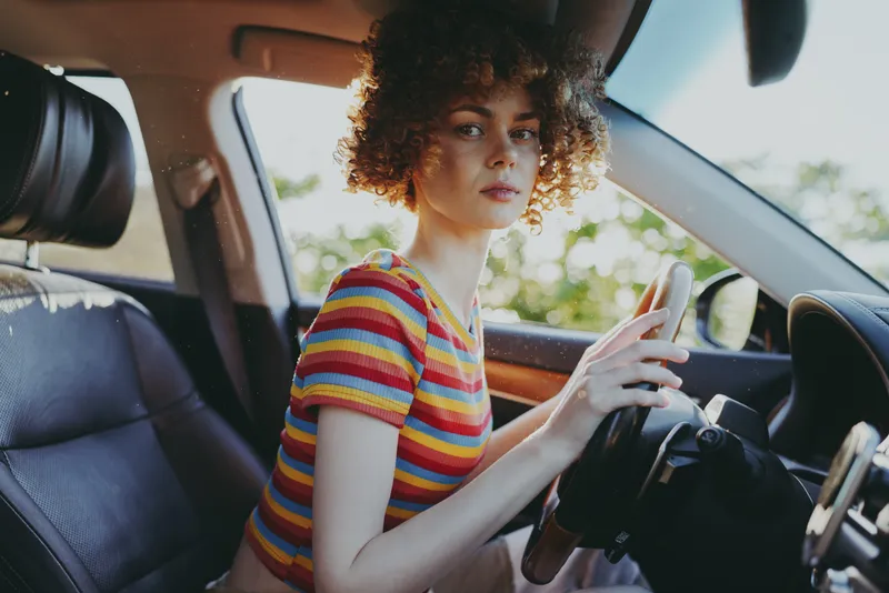 Young woman driver sits in a sunny car, gripping the wheel with focus as bright light pours through the window, creating a retro film vibe and a moment of everyday confidence.