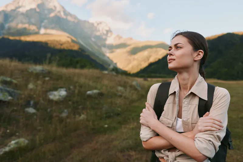 Woman lifestyle outdoors, casual hike, thoughtful gaze toward distant peaks, sunlit moment, relaxed posture, breeze through hair, natural colors, expansive mountain scenery.