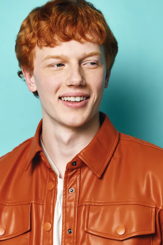 Young man with red hair and fair skin smiling and looking to side wearing brown leather jacket and white shirt on blue background in studio portrait.