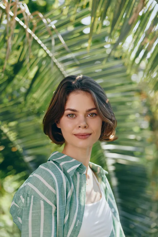 Portrait of a young woman outdoors among lush palm leaves, wearing a striped green shirt and white top, soft sunlight accentuates natural features.