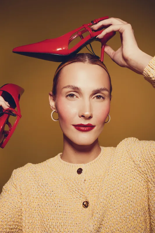 Young woman with red high heels holds shoes near head, wearing beige sweater and gold hoop earrings. Studio portrait with warm lighting and confident expression. Fashion and style concept.