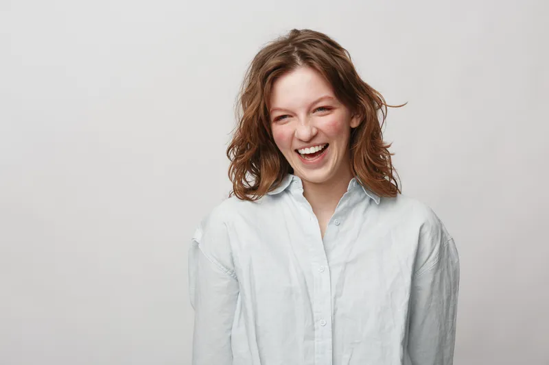 Happy young woman with curly hair laughing joyfully wearing a white shirt on light background. Concept of positive emotions, natural beauty, and casual lifestyle portrait.